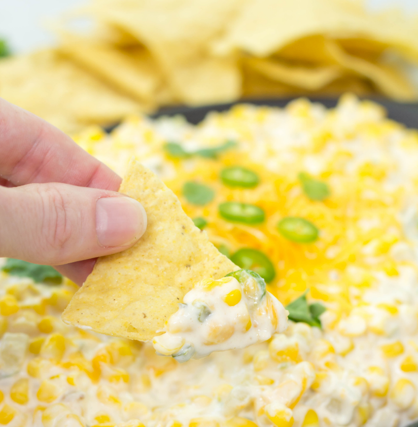 A person extends a chip into a cheesy dip with corn and jalapenos.