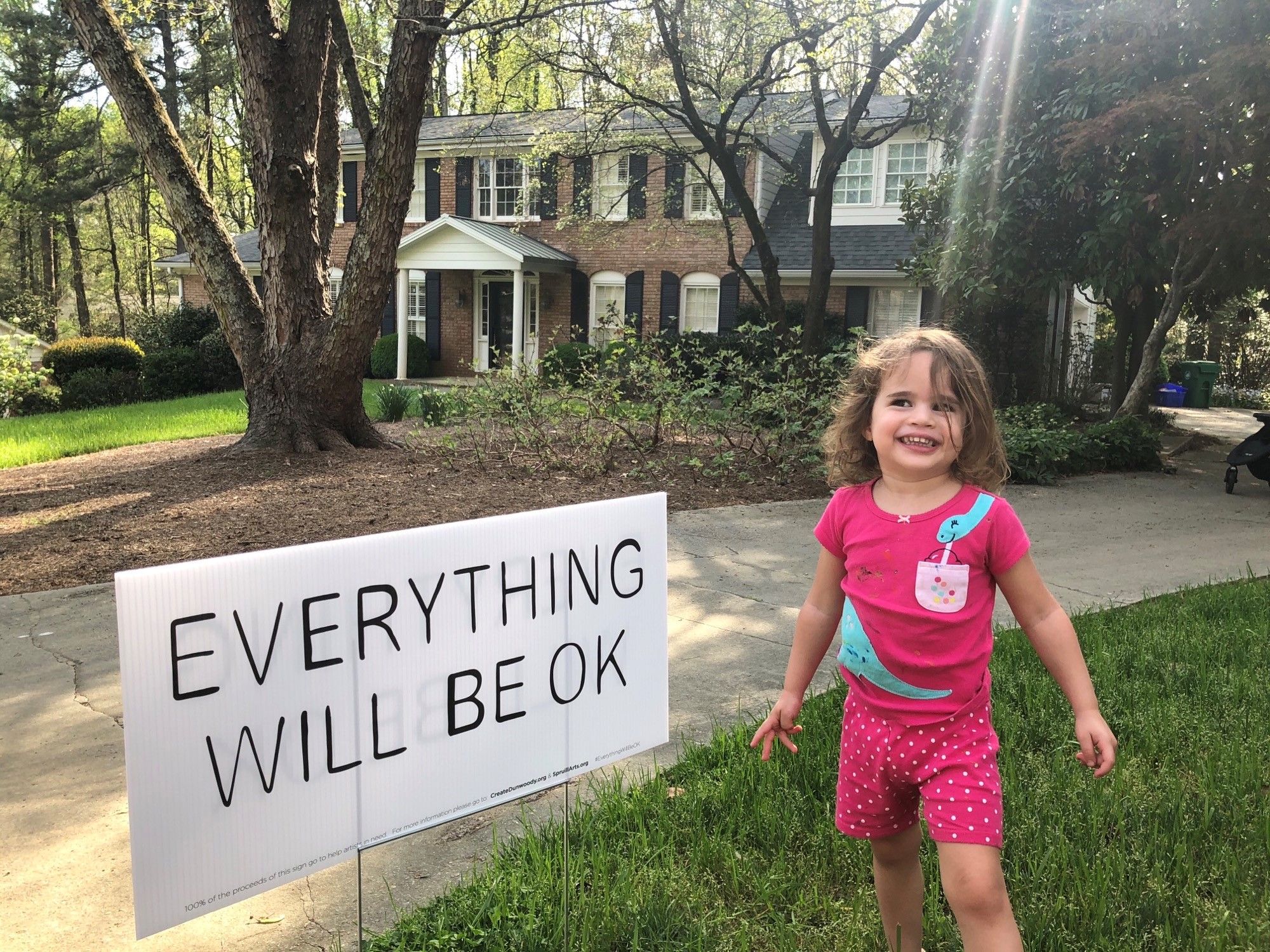 A female toddler stands next to a yard sign that says &quot;everything will be OK.&quot;