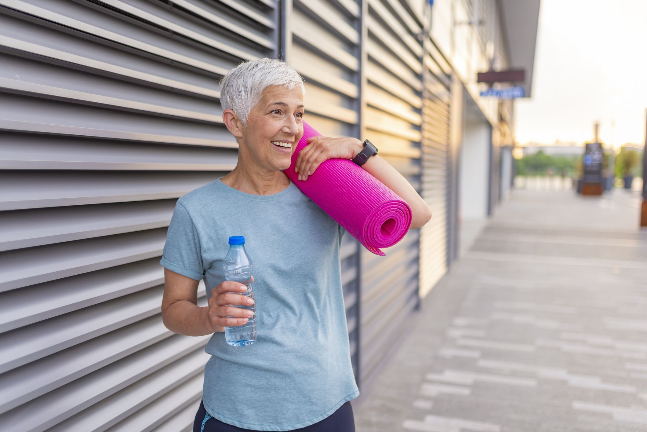 An older woman poses with a yoga mat and a bottle of water.